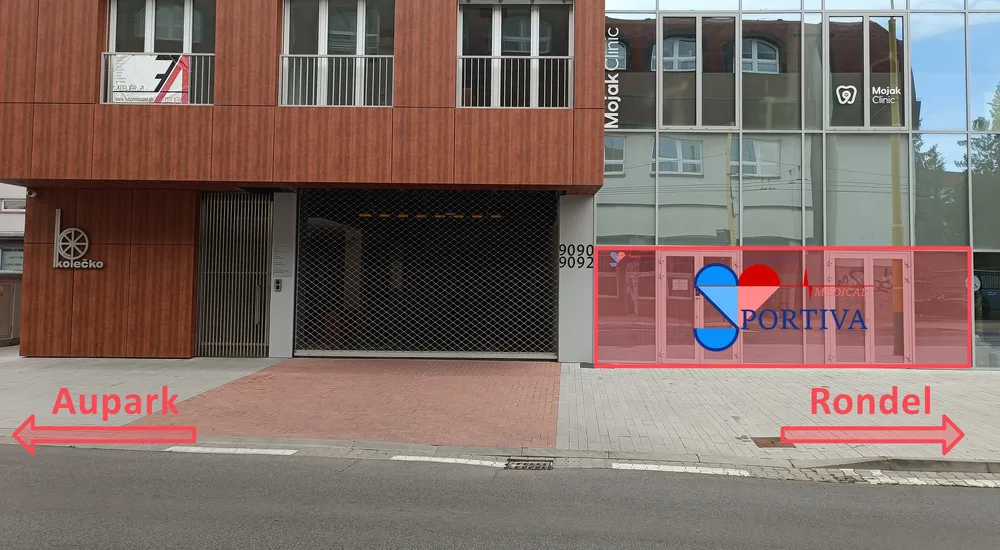 Priečelie budovy Residencia Kolečko z Hálkovej ulice. Vchod do ambulancie Sportiva Medical je zvýraznený červeným rámom, vpravo od vjazdu do garáže. Vľavo je smer na Aupark, vpravo na Rondel.